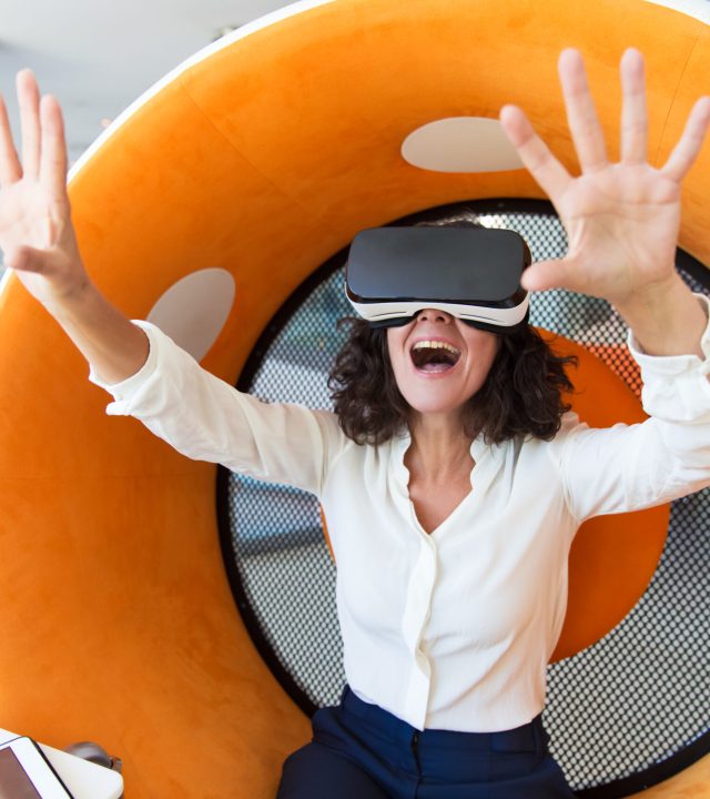 Excited businesswoman enjoying VR experience. Woman wearing virtual reality glasses, touching air, smiling and shouting. Future concept
